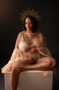 Pregnant Black woman in gorgeous shimmery gold bodysuit and crown, sitting upon a stool.