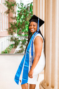 Senior woman graduating from MTSU wearing white dress, blue stole, and cords smiling at camera while leaning on pillar