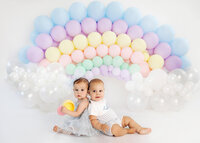 two babies sitting back to back with rainbow balloon arch during milestone portrait session in Burbank