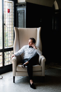 A refined composition of a groom in a tuxedo seated in a high-back leather armchair, reflecting an intentional presence and editorial style.