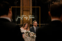 Mariés riants à la table d'honneur, enseigne au néon "just us two" sous une arche dorée au Château Vaudreuil, invités en silhouette au premier plan