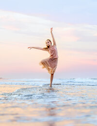 Young ballerina balancing in shallow ocean waves during a pastel sunset beach photography session with Nature’s Child Photography, soft pink skies and flowing skirt catching the coastal breeze as she poses gracefully in the surf.