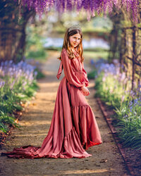 Child in a dusty pink gown standing beneath a blooming wisteria arch in a New Bern garden