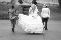 Black and white Venice Beach Los Angeles wedding photography of the bride and groom walking through the venue garden with a bridesmaid helping, captured in a candid documentary moment.