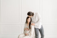 A maternity session taken at a Dallas/Fort Worth photography studio of an expecting mother sitting on a chair while her spouse is leaning over to kiss her forehead.