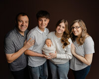 Family portrait with parents and siblings smiling while holding their newborn baby wrapped in a soft neutral blanket during a studio newborn photography session photographed by Kaitlyn Dawn Photography.