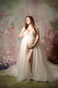 A pregnant woman in a tulle dress smiles up and out of the frame. She has one arm bent against her chest with her hand in her hair while the other arm drapes around her bump, holding it from beneath. She stands in front a multi-colored floral backdrop.