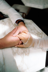 Bride and groom holding hands across a marble table, with custom embroidered wedding gloves reading “Aug 2024,” captured during Japna and Chris’s Hotel Chelsea elopement in New York City by wedding photographer Perry Hancock.