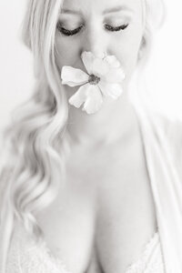 Light filled black and white boudoir photo of a woman wearing lace lingerie while she has a fresh flower in her mouth.