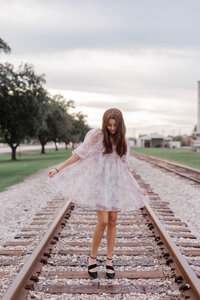 Girl of color in Katy Texas  looks down at her black heels while wearing a pink floral dress standing on railroad tracks