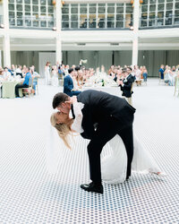 Groom dips his bride during their first dance in a bright circular ballroom as guests look on from surrounding tables at Dayton Arcade wedding.