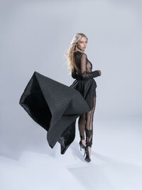 Fashion portrait of a model in a sheer black gown holding a large angular sculptural fabric form against a pale backdrop.Woman in a cream blazer and black trousers leaning against a white wall beside a window, one hand on her hip.