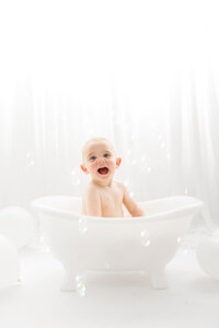 Baby smiles at the camera while sitting in a miniature claw foot splash bath tub during his first birthday Cake Smash Photoshoot at Holy City Babies Portrait Studio in Charleston, SC.