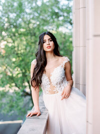 bride standing on a balcony with greenery in the background and soft daylight highlighting her gown