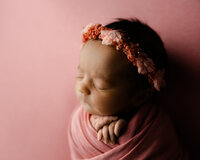 Sleeping newborn baby girl wearing a pink floral headband and wrapped in a soft pink swaddle on a blush backdrop during a studio newborn photography session in Yucaipa by Kaitlyn Dawn Photography.