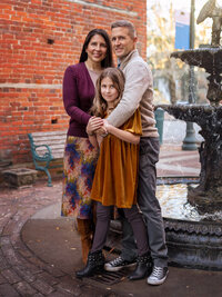 Parents and daughter posing together near a brick wall and fountain during a downtown family portrait session