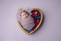 Sleeping newborn baby girl wrapped in a lavender swaddle and posed in a heart-shaped bowl prop during a creative studio session with newborn photographer Kaitlyn Dawn Photography.