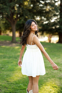 High school senior girl walking through a grassy park wearing a white dress during golden hour, senior portrait by Kaitlyn Dawn Photography.
