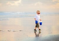 Young boy in red sunglasses carrying two blue buckets while walking along the shoreline on reflective wet sand during a beach child Beach. - A portrait experience by Nature's Child Photography.