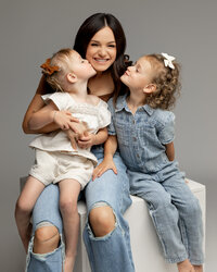 Mother sitting on a studio cube with her two young daughters laughing and cuddling, soft gray backdrop and natural tones photographed by Kaitlyn Dawn Photography, studio family portrait specialist.