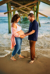 Pregnant couple standing beneath a pier during a beach maternity session in Atlantic Beach North Carolina