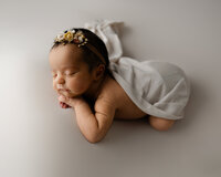 Newborn baby girl peacefully sleeping while wearing a floral headband and white wrap on a soft neutral backdrop during a professional newborn photography session by Kaitlyn Dawn Photography in Yucaipa.