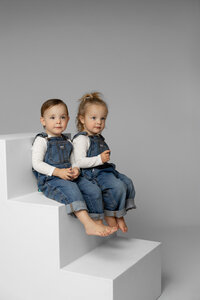 Two young siblings sitting side by side on white studio steps, coordinated denim outfits photographed by Kaitlyn Dawn Photography, sibling portrait photographer.