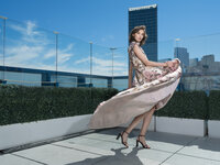 Model in a floral gown with a long train lifting in the wind on a rooftop terrace above the city.