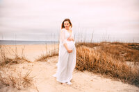 A pregnant woman stands between sand dunes on a golden beach. She stares off while holding her hands above and below her belly.
