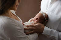 Close-up lifestyle portrait of a newborn baby sleeping in their parents’ arms while wearing a delicate headband during an intimate newborn photography session by Kaitlyn Dawn Photography.
