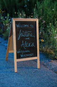 A chalkboard sign sits by a garden path, reading “Welcome to Justine and Alex’s Wedding Weekend!” in handwritten script.