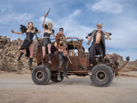 Costumed performers posed on a rusted desert vehicle with arms raised against an open sky.