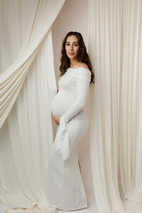 Pregnant woman in a fitted white gown gently holding her belly, standing between soft draped fabric during a maternity session with Kaitlyn Dawn Photography in Yucaipa.