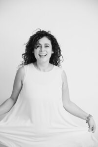 A black and white picture of a woman with dark curly hair smiling softly down, standing on a white backdrop. She wears a white dress and holds it loosely away from her body.