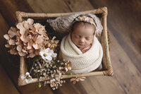Newborn baby girl wrapped in a white hand-knitted wrap, posed in a wicker crib at Jennifer Brandes Photography near Mankato, MN.