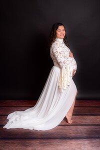A pregnant woman in a flowing white dress smiles as she poses for a photo, showcasing her beautiful baby bump.