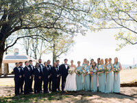 Bride and groom standing with their full wedding party, bridesmaids in light blue dresses and groomsmen in black tuxedos, on the lawn at Burritt on the Mountain in Huntsville, Alabama, photographed by Perry Hancock.