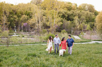 A family of five and their loyal dog stroll through a grassy field in a Toronto park, surrounded by lush trees and greenery under a clear sky, capturing the perfect moment for family photography.
