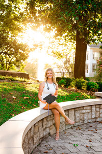 Senior holding graduation cat sitting on the bench at Trevecca University with golden light shining through the fountain beside the bell tower