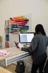 Woman with dark hair working on a laptop beneath shelves of colorful embroidery threads. Photograph by Yucaipa branding photographer Kaitlyn Dawn Photography.