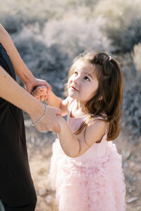 Young daughter from the Parker family holding her mother’s hands and looking up at her during a warm, sunlit family photography session in the Las Vegas desert.