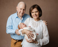 Grandparents smiling while holding their newborn grandchild wrapped in a soft neutral swaddle during a studio newborn family portrait by Kaitlyn Dawn Photography.