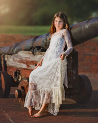 Child in white lace dress seated on historic cannon during golden hour portrait session in Eastern North Carolina
