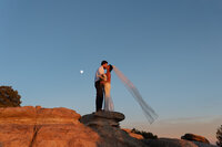 Bride and groom kissing atop a red rock formation at dusk, her veil flowing in the wind