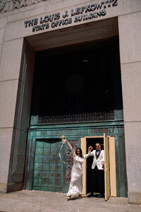 Newlyweds celebrate outside New York City Hall, holding hands and raising a bouquet in joy as they step through the grand bronze doors.