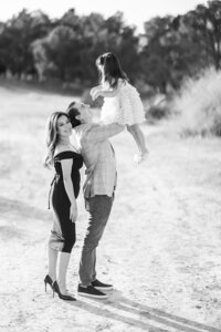 Black and white image of the Parker family in Las Vegas, with a father lifting his young daughter in the air while the mother smiles beside them during a joyful outdoor family photography session.
