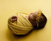 Sleeping newborn baby girl wrapped in a mustard yellow swaddle and wearing a floral headband on a yellow backdrop during a studio newborn photography session.
