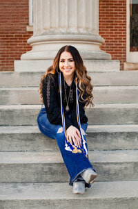 Senior girl wearing jeans and black top smiling at camera while sitting on the MTSU stairs of campus building