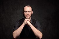 A dark haired man with light blue eyes stares ahead seriously with his hands folded under her chin. He wears a gray shirt with a black vest and sits in front of a gray backdrop. The lighting is dramatic.