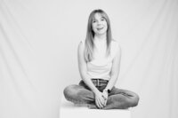 A black and white picture of a blonde woman with straight hair sitting, cross-legged on a white box in front of a white backdrop. She wears a white shirt and jeans. Her hands rest softly in front of her lap, and she appears to laugh gently.